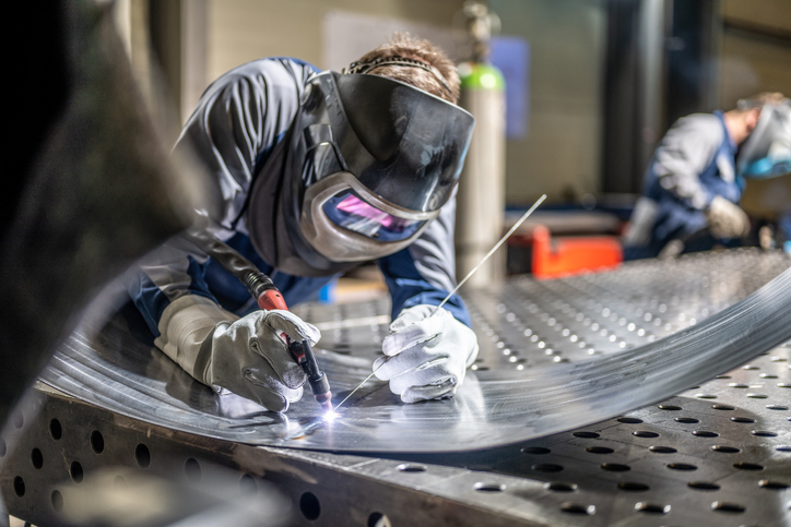 Male welder wearing helmet working with welding torch in factory, practicing common sheet metal fabrication techniques.
